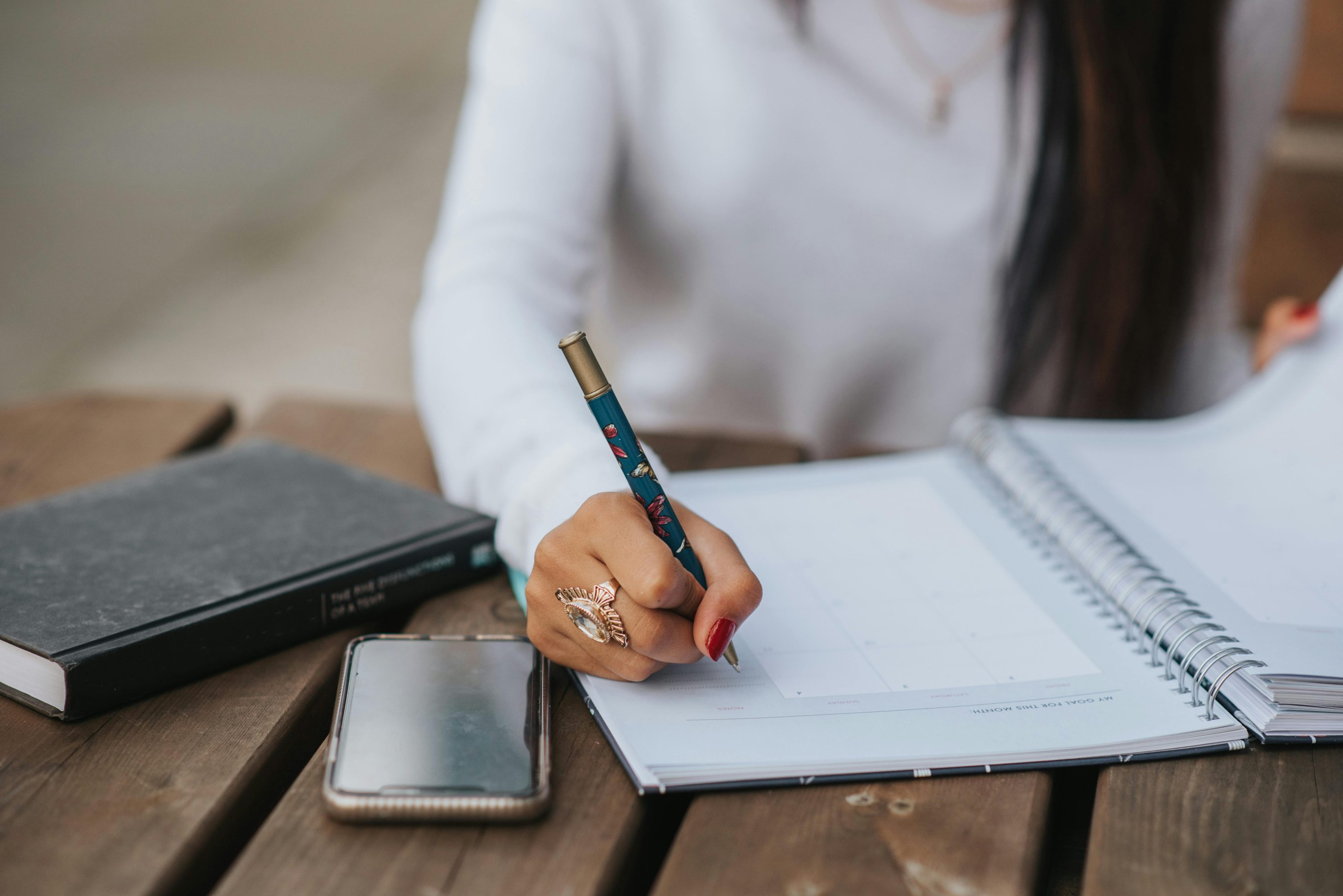 Crop unrecognizable female worker writing with pen in notepad at table with smartphone and diary