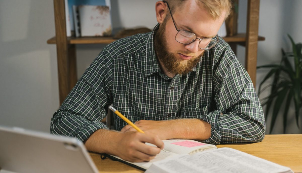 Man studying and taking notes with a Bible on a desk indoors.